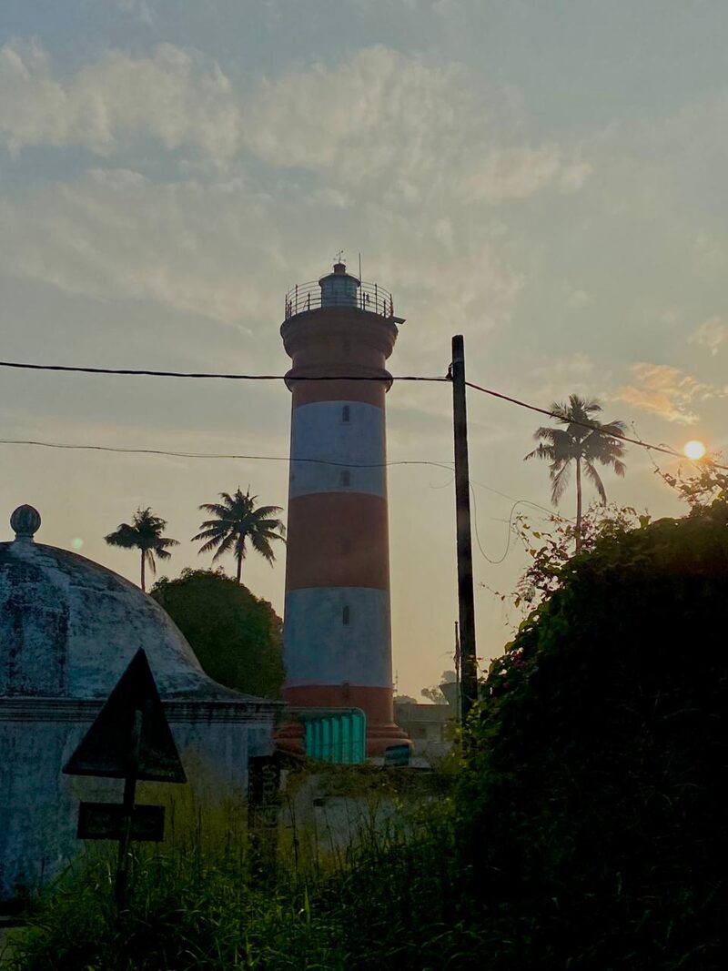 Red and white striped lighthouse at first light with palms and a small shrine nearby