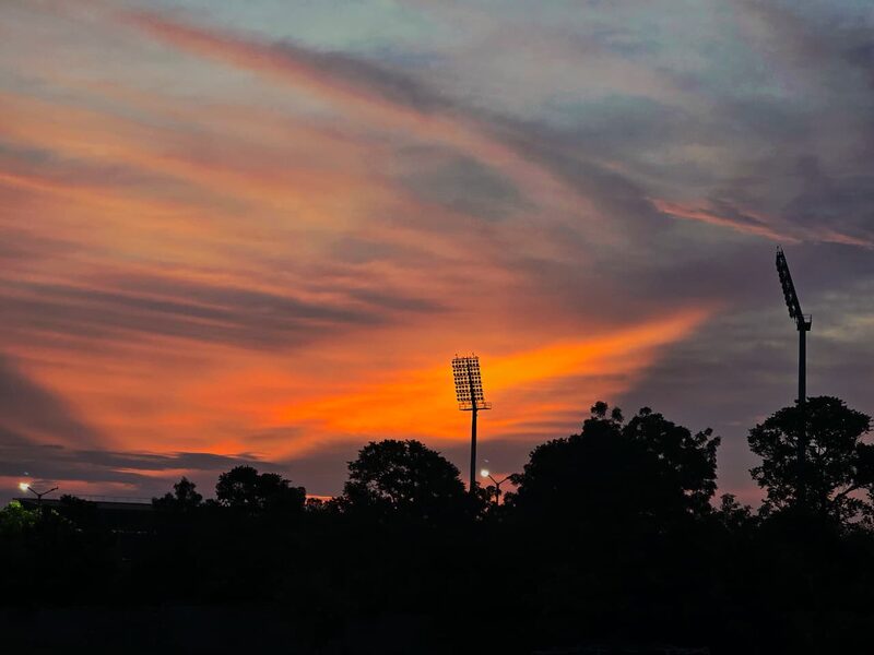 Orange and violet dusk sky behind silhouetted stadium floodlights and trees
